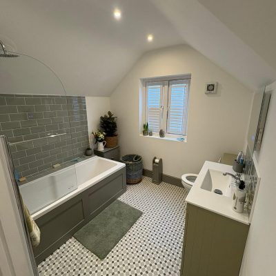 Modern loft bathroom with sloped ceiling, grey bath and vanity, green tiled shower wall, patterned floor, and window with white shutters. iGroup South West Ltd.